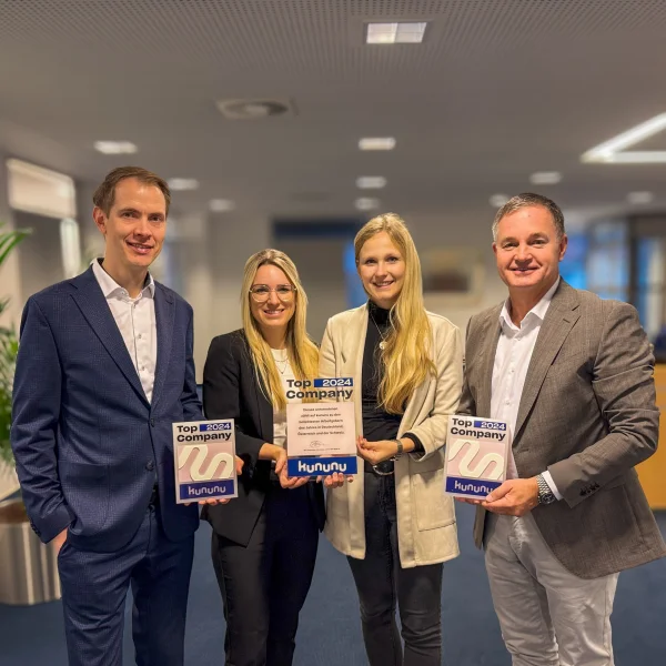 Kununu Top Award Auszeichnung 2024: Vier Personen (von links nach rechts: Matthias Smit, Larissa Lorenz, Janina Köster, Matthias Kruse) stehen nebeneinander in einem modernen Büroinnenraum und halten jeweils eine „kununu Top Company 2024“-Auszeichnung in den Händen. Die Gruppe ist frontal zur Kamera ausgerichtet, im Hintergrund sind unscharf Büromöbel, Deckenleuchten und Pflanzen zu erkennen.